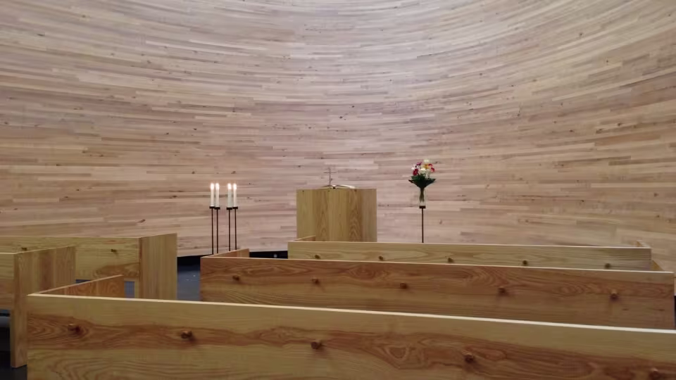 Im Inneren der Kapelle des Schweigens herrscht eine warme, friedliche Atmosphäre mit Holzbänken, vier brennenden Kerzen und einem schlichten Altar mit einem Blumenarrangement.